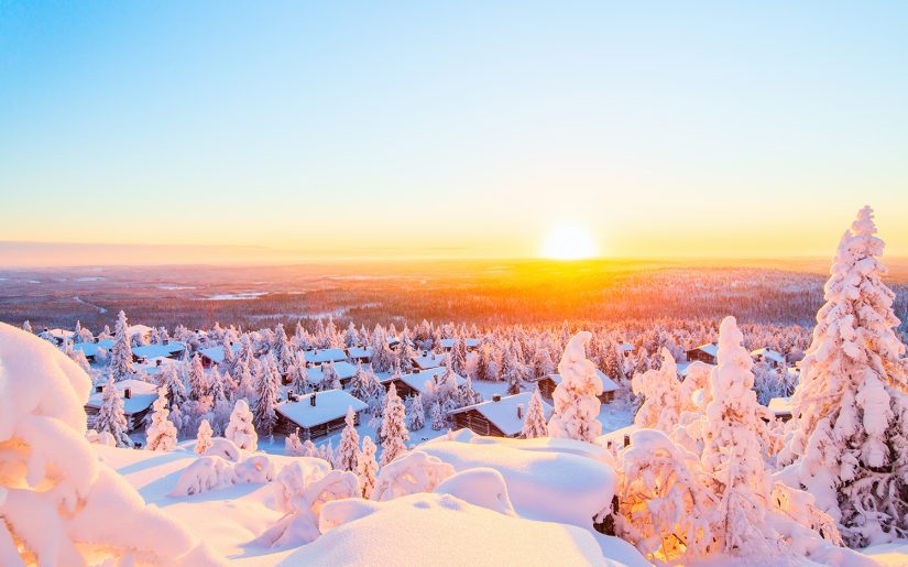 Eine Panorama Aussicht über Finnlands Lappland. Verschneite Wälder sowie ein Dorf im Vordergrund. Sonnenaufgang